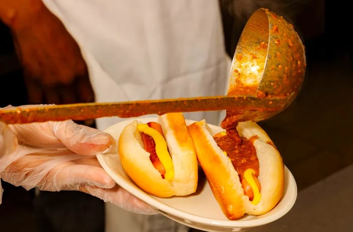 A ladle pours chili over two coney hot dogs already covered in mustard.