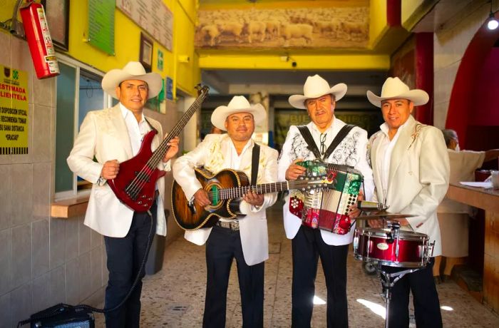 A four-member band, featuring bass, guitar, accordion, and drums, stands in a spacious hallway. They are dressed in decorative suit jackets and cowboy hats.