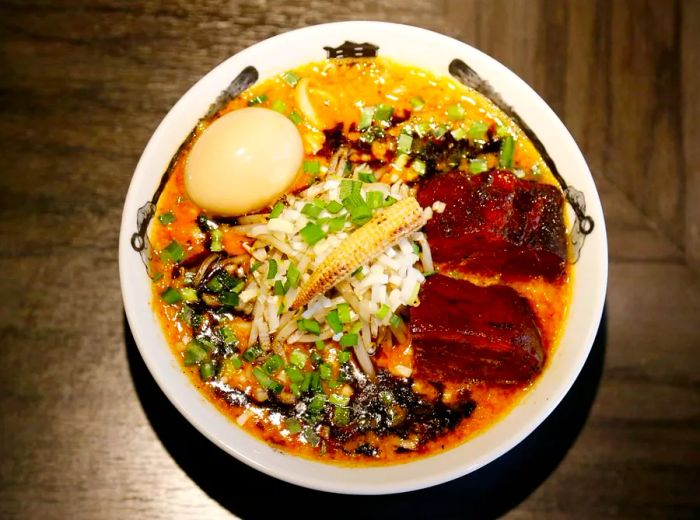 From a bird's-eye view, a bowl brimming with toppings such as caramelized pork slices, green onions, a boiled egg, baby corn, and swirls of dark spice mix.