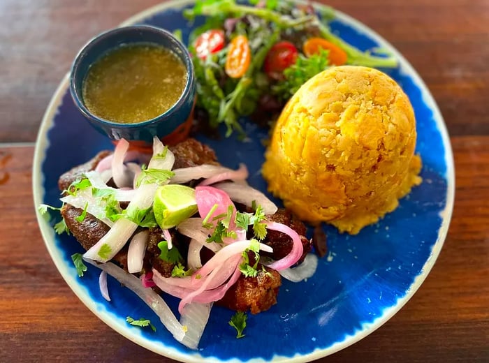 A vibrant mound of mofongo alongside pork topped with pickled onions, a fresh salad, and a side of green sauce.