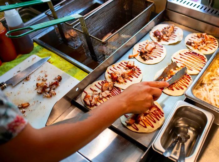 A chef uses tongs to position meat on a series of tortillas laid out on a plancha, all already drizzled with sauce.