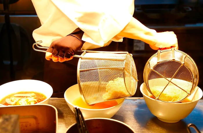 A chef folds his arms while serving noodles into two bowls of ramen.