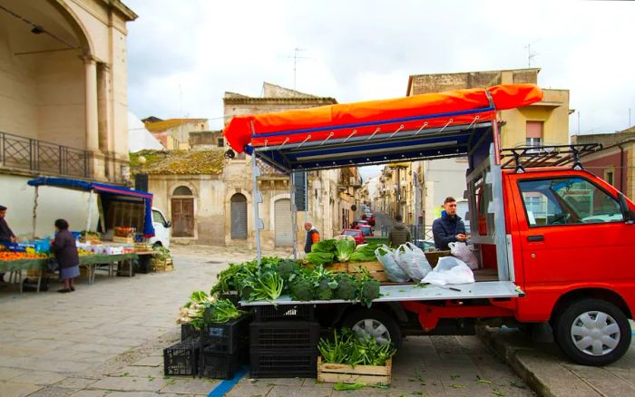 At an open-air farmer’s market in central Comiso, Sicily, a senior customer shops among the vibrant fruit and vegetable stalls even in winter.