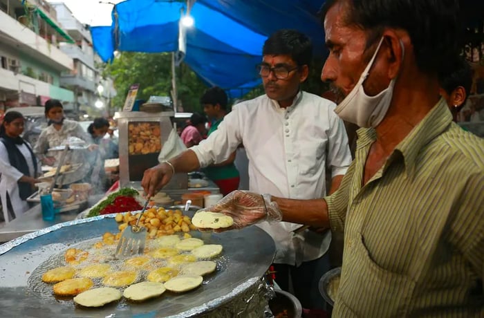 Cooks prepare aloo tikki on a large tawa at a street stall, with customers gathered around.