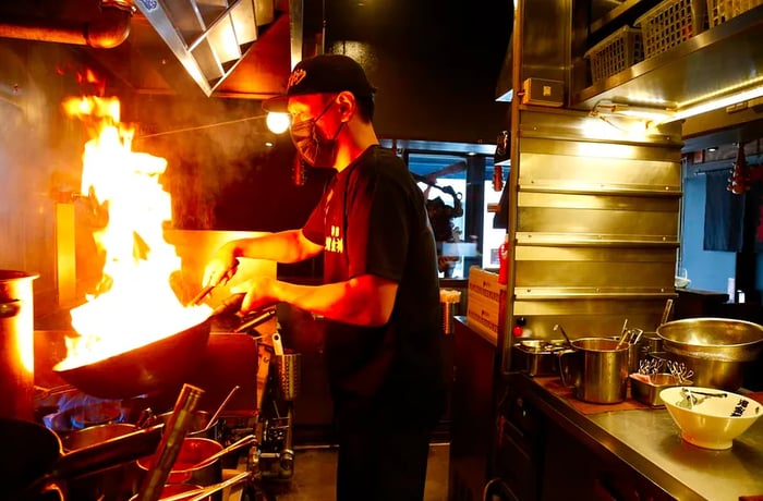 A chef operates a wok, surrounded by a dramatic burst of flames.