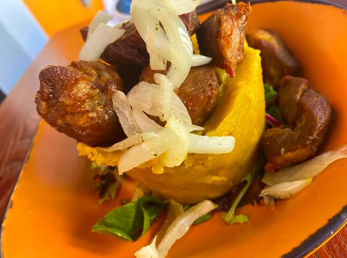 A shallow ceramic bowl filled with mofongo, generously topped with large pieces of meat and sliced onions.