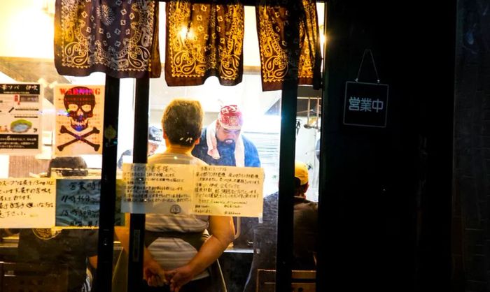 A chef is seen through a dimly lit window, hard at work behind the ramen counter.