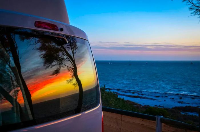 A camper van parked by the ocean at sunset, with the vibrant sky and trees mirrored in its rear window.