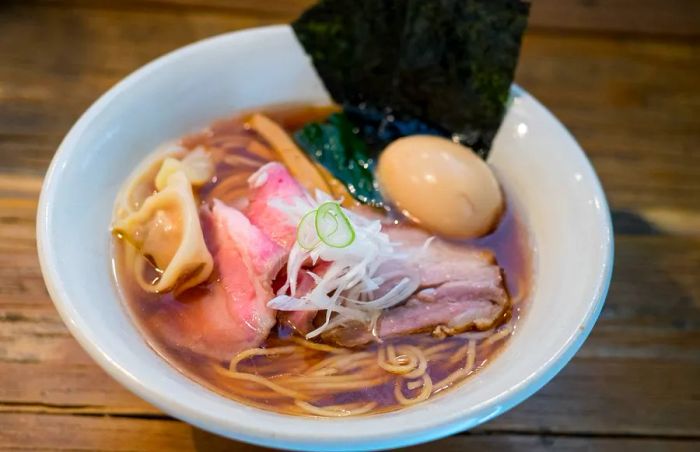 A bowl of ramen features relatively thin noodles topped with layers of pork, a wonton, bamboo shoots, a slice of seaweed, and a boiled egg floating atop the broth.