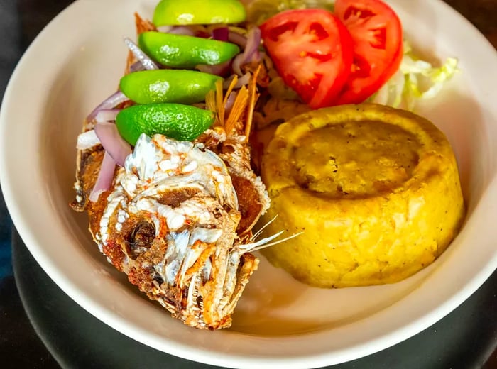 A serving of mofongo next to a whole fried fish garnished with lime wedges and accompanied by a salad.