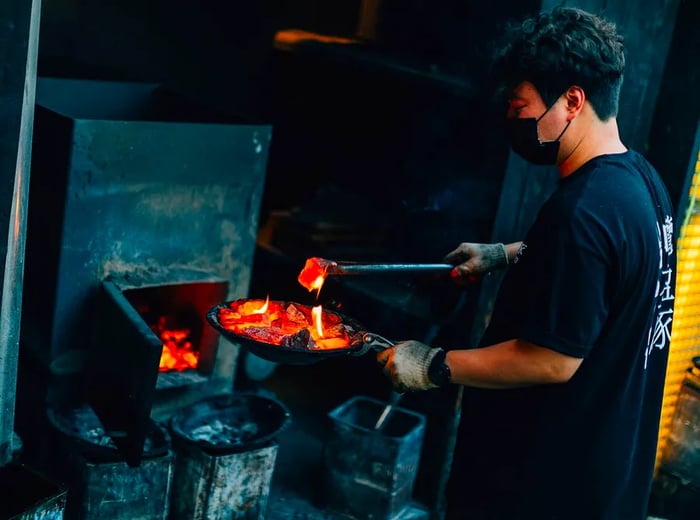 An employee uses tongs to transfer a hot coal from a pan to a furnace.