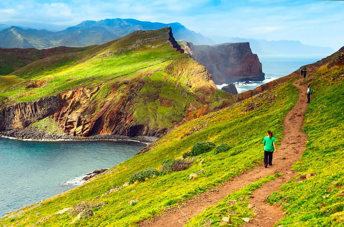 Three hikers traverse a grassy coastal path, one gazing down at the sea, with rugged mountains rising in the background.