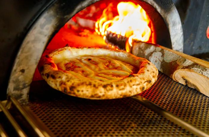 A pizza on a metal paddle being slid into an oven ablaze with large flames.