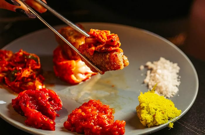 Chopsticks lift a piece of grilled meat topped with kimchi, resting on a plate surrounded by various banchan.