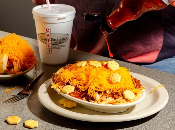 A diner adds hot sauce to a plate of spaghetti topped with chili and cheese. Next to it sits a carney (hot dog) heaped with cheese, accompanied by a styrofoam cup from Camp Washington Chili.