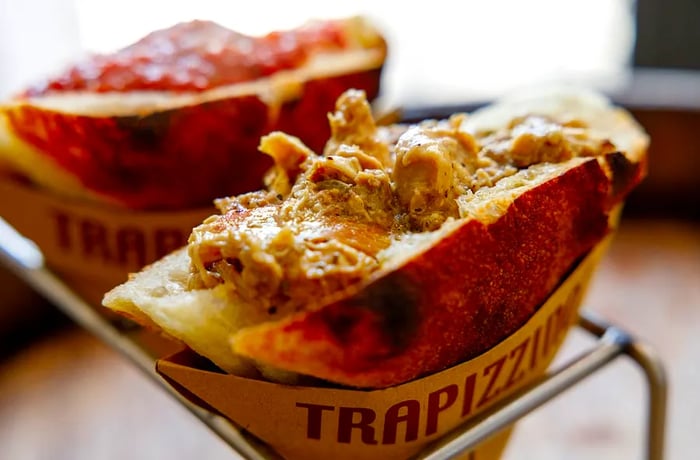 A pizza pocket filled with stewed chicken, resting in a unique metal holder with a cardboard sleeve labeled Trapizzino.