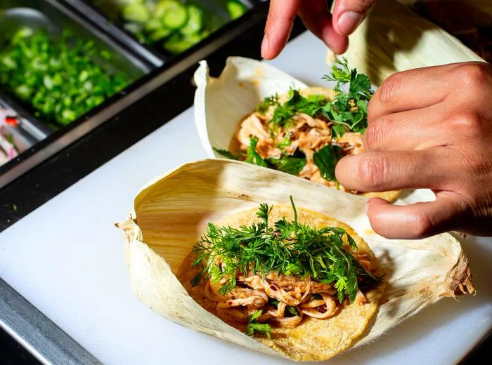 Hands craft two tacos on a prep counter, filled with shredded meat and garnished with cilantro.