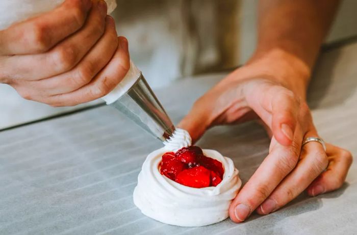 A pastry chef skillfully uses a piping bag to swirl decorative cream around a center of cooked strawberries.