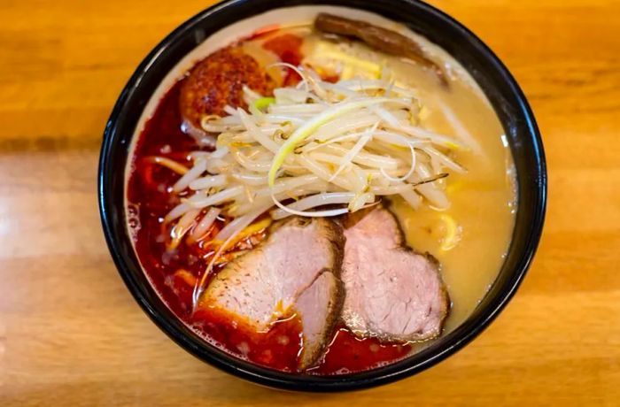 A bird's-eye view of a ramen bowl featuring two thick slices of pork, sprouts, and a contrasting deep red broth on one side with a pale yellow on the other, all set on a wooden table.