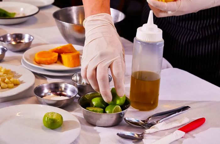 A gloved hand reaches for a bowl of limes resting on a tablecloth-covered surface, where various ingredients are arranged.