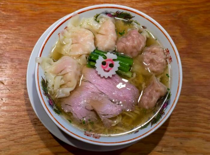 A beautifully arranged bowl of ramen sits on a wooden counter, featuring tender slices of pork and two types of wontons.