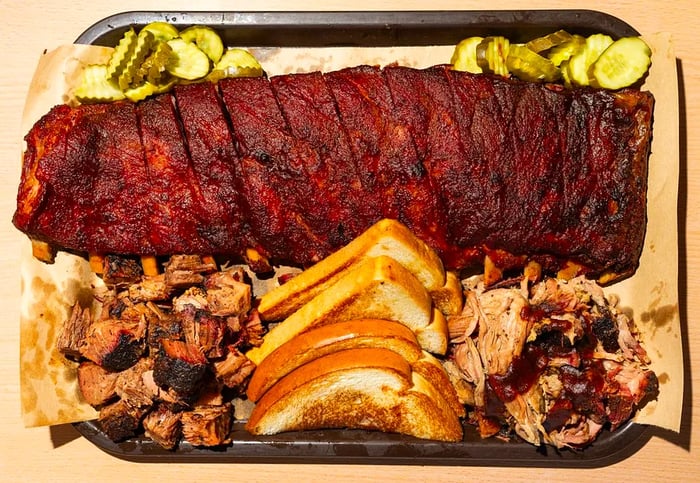 From above, a platter of grilled meats featuring a full rack of ribs, burnt ends, and pulled pork, accompanied by slices of toasted white bread.