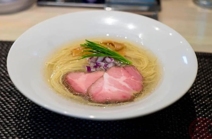 A spacious bowl of ramen featuring noodles and pink slices of pork floating in a light straw-colored broth.