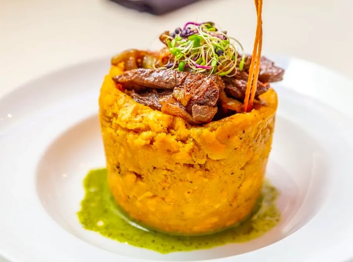 A tall mofongo, adorned with thick slices of steak, sprouts, and various garnishes, resting in a pool of green sauce.