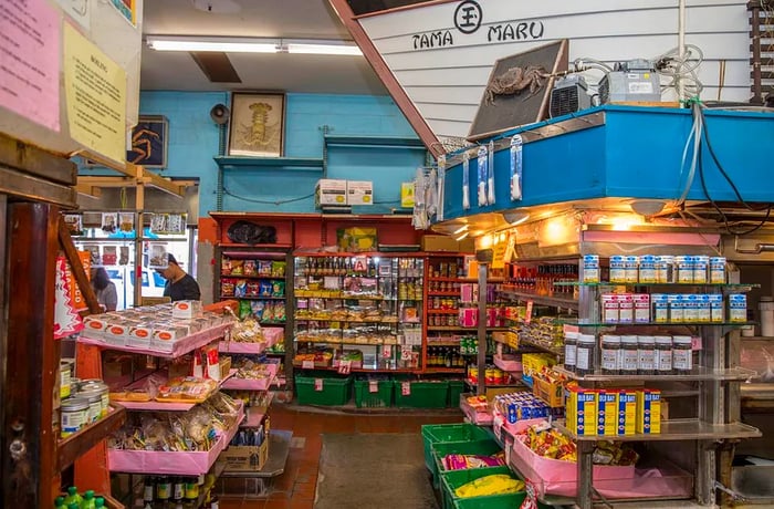 The interior of a grocery store showcases product shelves beneath a full-sized boat hull, adorned with the words ‘Tama Maru’.