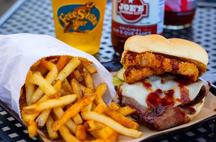 A brisket sandwich adorned with cheese, barbecue sauce, and onion rings on a thick bun, accompanied by a scattering of fries on a metal serving tray.