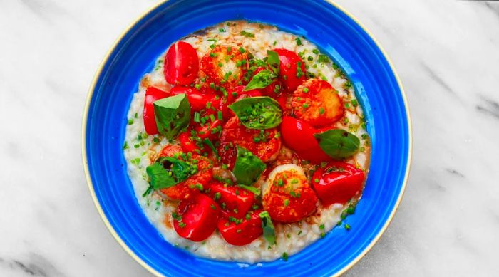 An aerial view of seared scallops, vibrant red tomatoes, and basil leaves atop a creamy risotto in a blue bowl.