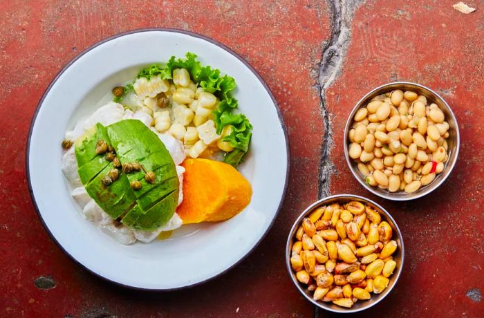 An overhead view of a ceviche bowl being prepared, topped with slices of avocado and capers over a mound of fish, accompanied by a chunk of cooked sweet potato and bowls of corn nuts.