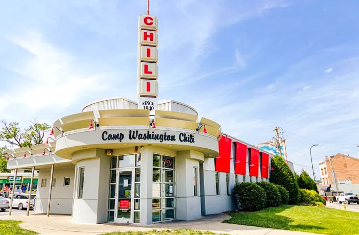 The view of a restaurant from the street corner features a towering spire labeled ‘chili’ rising from the roof. Cursive writing above the door states Camp Washington Chili.