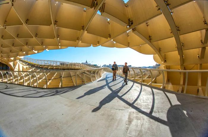 Two individuals stroll along the famous Las Setas (or Metropol Parasol) in Seville, Spain, beneath a clear blue sky.