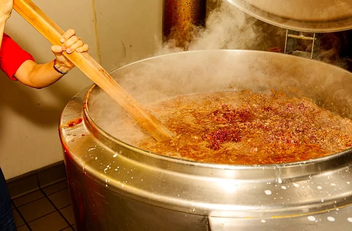 A staff member stirs a large pot of chili with a long wooden spoon.