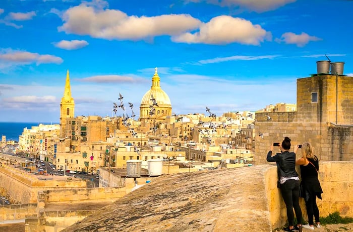 Two female tourists capture a photo of pigeons with the stunning skyline of Malta's capital, Valletta, featuring its dome and bell tower under clear blue skies.