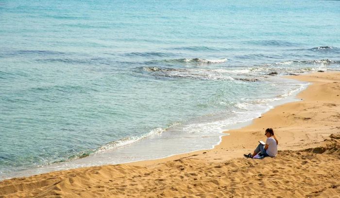 A woman enjoys reading a book on the sandy beach during winter in Protaras, Cyprus.