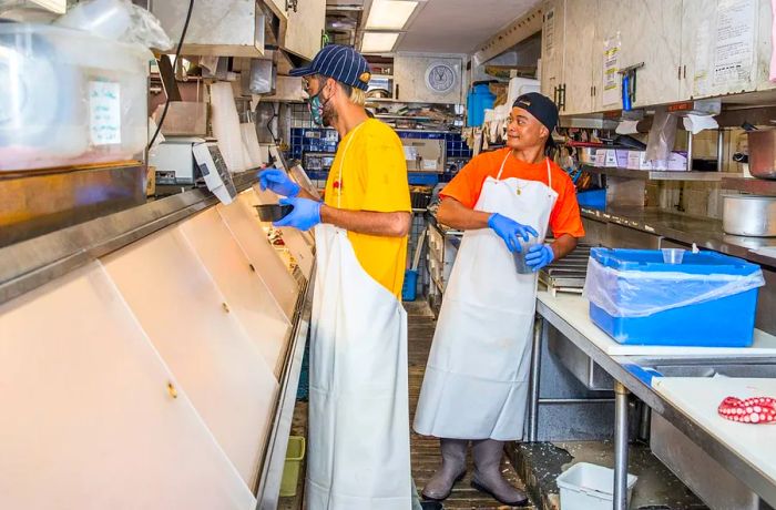 In a prep area behind the deli counter, two workers converse with a customer.