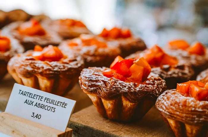 Layered tartlets adorned with apricot slices are displayed, accompanied by a small sign in French indicating the price: 3,40.