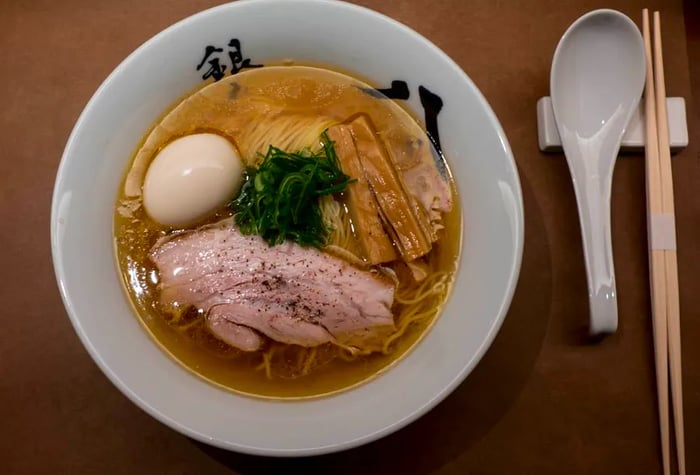 From above, a bowl of ramen showcases thick pork slices, bamboo shoots, a boiled egg, fresh greens, and noodles, with chopsticks and a spoon resting on the counter.