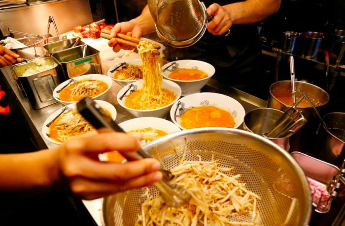 In a bustling prep area, workers add noodles and toppings to bowls filled with noodles.