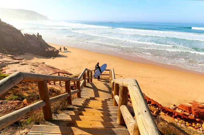 A surfer makes their way down a wooden staircase to Praia do Amado, located near Sagres in the Algarve, Portugal.