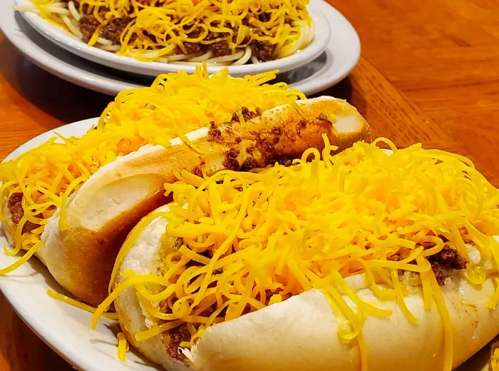 A plate featuring two coneys generously topped with cheese alongside a serving of three-way spaghetti.