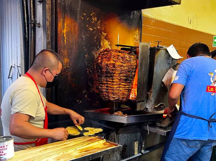 Two chefs skillfully maneuver around a trompo, charring meat over an open flame in the kitchen.