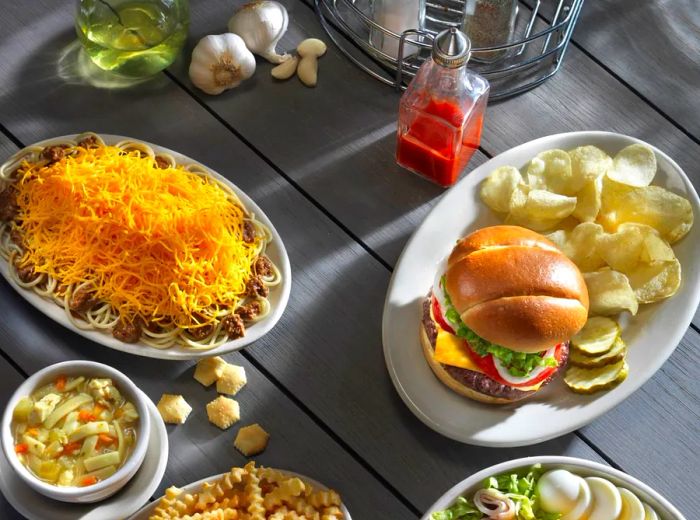 An aerial view of a picnic table spread with dishes featuring chili and cheese over spaghetti, a cheeseburger, onion rings, fries, and a chef’s salad.