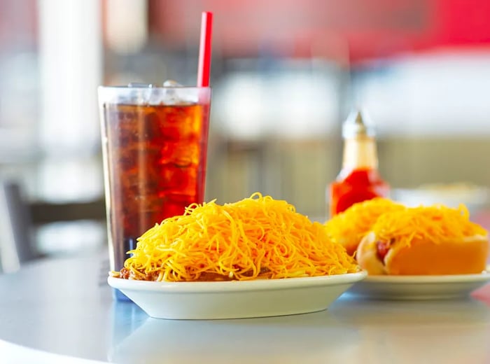 A plate featuring spaghetti topped with chili and cheese, alongside coneys and a tall soda.