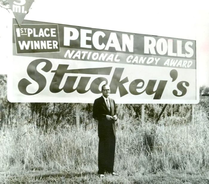 A black and white photograph captures W.S. “Sylvester” Stuckey, Sr. standing before a roadside billboard proclaiming, “First place winner. Pecan Rolls. National Candy Award. Stuckey’s.”