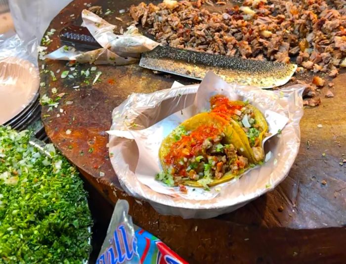 Two tacos drenched in salsa sit on a paper plate next to a comal, accompanied by a large chef’s knife and chopped meat.