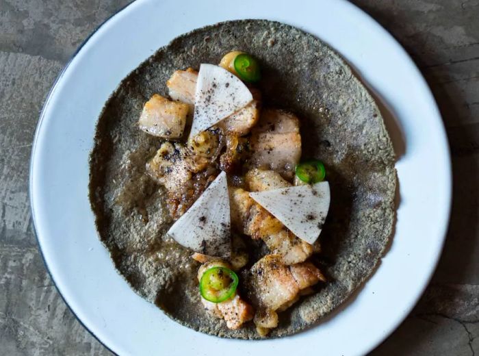 An overhead view of a dark tortilla on a plate, featuring slices of jalapeño, radish, and fish drizzled with a rich sauce.