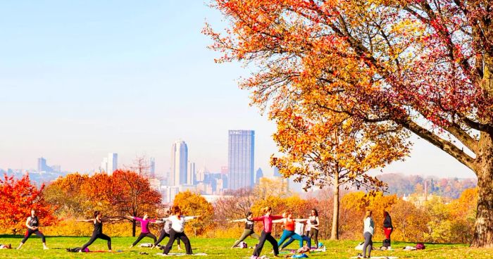 A group of women participating in a yoga class at Schenley Park, with the hazy Pittsburgh skyline in the background on a fall morning